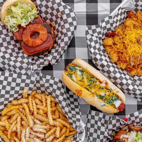 A variety of fast food items on checkered paper: a burger with pineapple, hot dogs, tater tots, and crinkle-cut fries.