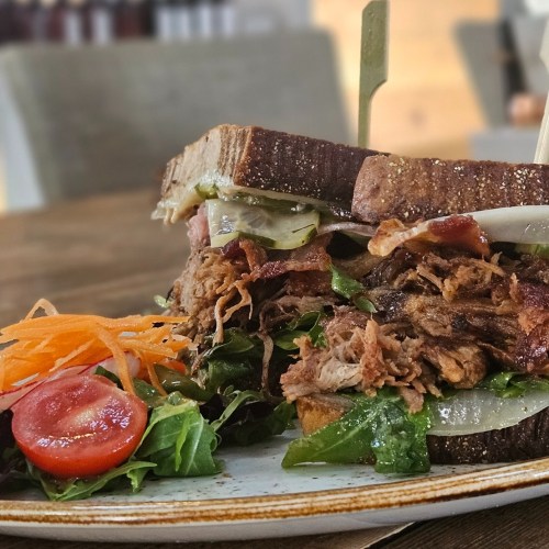 A close-up of a pulled pork sandwich with lettuce, pickles, and a side salad on a wooden table.