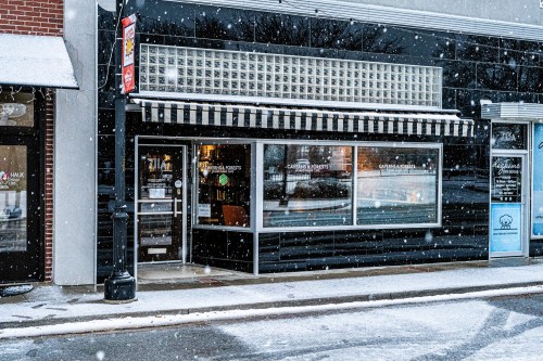 A storefront with large windows and a striped awning, surrounded by falling snow on a quiet street.