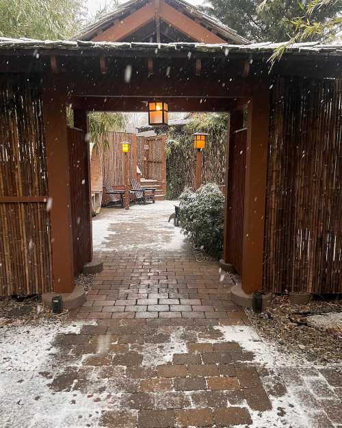 A snowy pathway leads through a wooden archway into a serene garden with bamboo and warm lanterns.