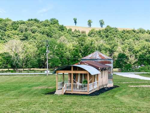 A small, rustic house with a metal roof and porch, surrounded by green fields and trees under a clear blue sky.