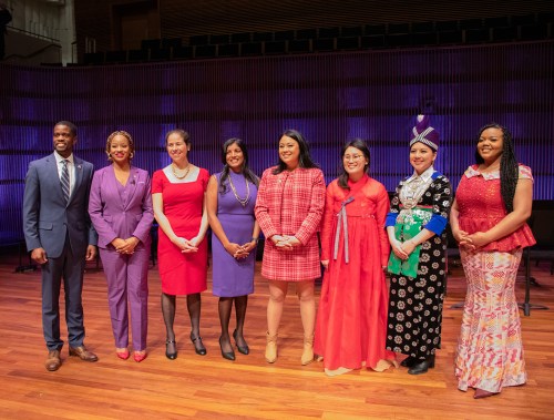 A diverse group of eight women and men in formal attire, standing together on a stage with colorful lighting.