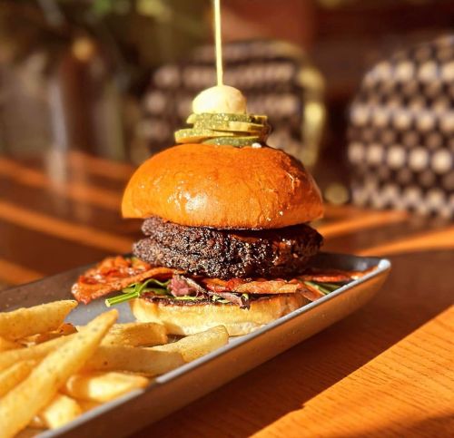 A gourmet burger with bacon and greens, topped with pickles, served with a side of golden fries on a metal plate.