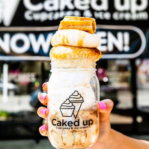 A hand holds a jar filled with ice cream, topped with a cupcake and drizzled with caramel, against a shop backdrop.