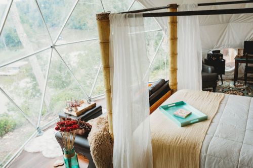 Cozy bedroom in a geodesic dome, featuring a four-poster bed, natural light, and a view of greenery outside.