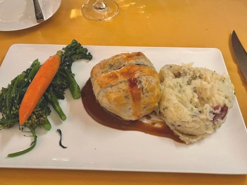 A plate featuring a pastry-wrapped dish, creamy mashed potatoes, a carrot, and broccolini drizzled with sauce.