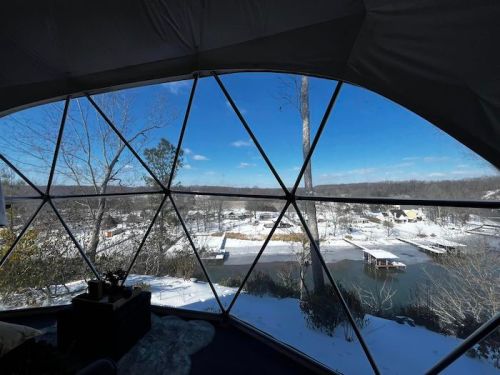 View from a geodesic dome window overlooking a snowy landscape and a calm river with boats. Clear blue sky above.