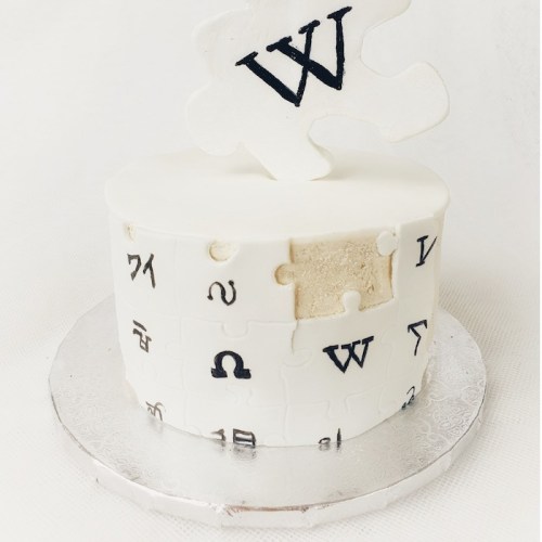 A round white cake decorated with puzzle pieces and a large "W" on top, featuring various letters and symbols.