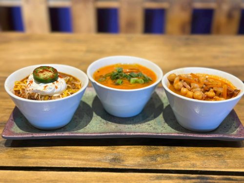 Three bowls of colorful dishes on a green plate: one with noodles, one with a creamy soup, and one with chickpeas and vegetables.