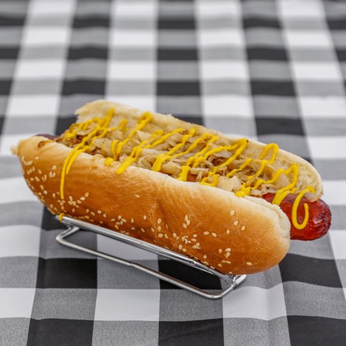 A hot dog in a sesame seed bun topped with mustard and sauerkraut, served on a silver holder against a checkered tablecloth.