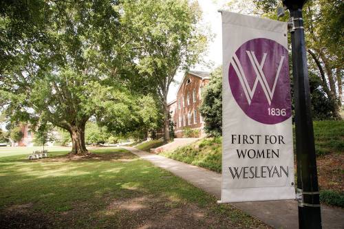 A banner reading "First for Women Wesleyan" stands in a green area with trees and a brick building in the background.