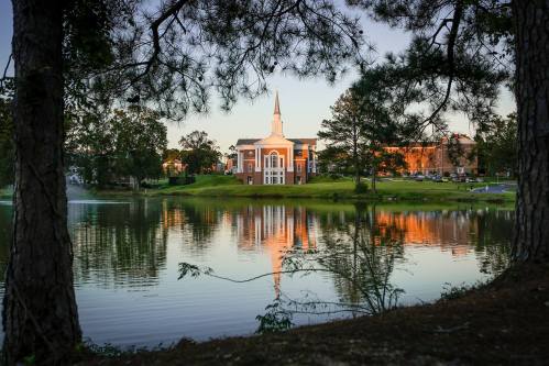 A serene lake reflects a church with a steeple, framed by trees at sunset.