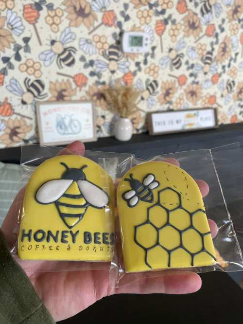 Two yellow cookies shaped like bees and honeycomb, packaged and held in a hand, with a floral background.