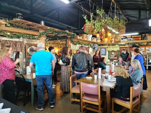 A lively restaurant scene with people chatting and dining at tables, surrounded by rustic decor and hanging plants.