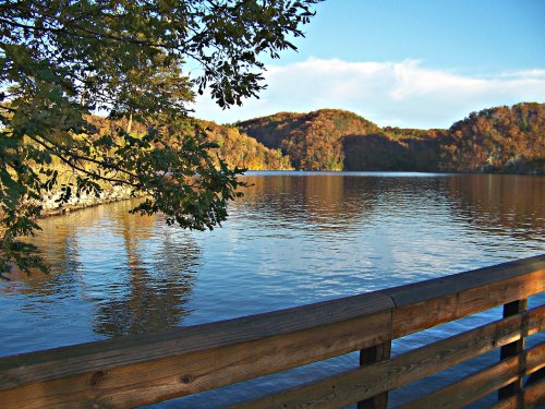A serene lake surrounded by autumn-colored hills, with a wooden railing in the foreground and clear blue skies.