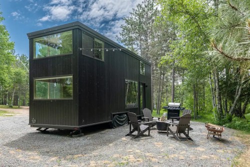 A modern black tiny house with large windows, surrounded by trees, featuring a fire pit and grill in a gravel area.