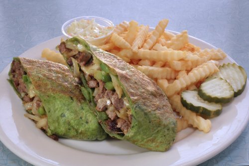 A plate featuring two green wraps filled with beef and vegetables, served with crinkle-cut fries, pickles, and coleslaw.