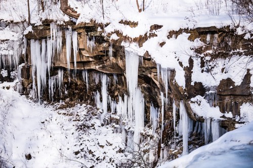 Icicles hang from a snowy cliffside, surrounded by a winter landscape of snow and rocky terrain.