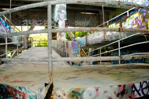 Abandoned concrete structure with graffiti-covered walls and a rusted railing, surrounded by greenery.
