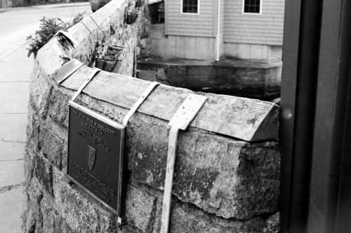 A stone wall with a plaque attached, beside a building, captured in black and white.
