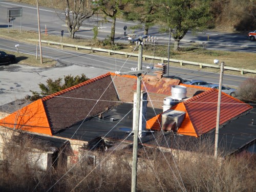 Aerial view of a building with orange and brown roofs, surrounded by power lines and a road in the background.