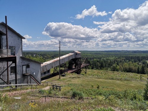An old industrial building with a conveyor belt overlooks a lush green landscape under a cloudy sky.