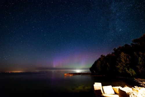 A serene night scene featuring a starry sky, faint auroras, and a calm lake with a wooden pier and trees along the shore.
