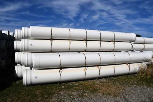 Stacked white pipes resting on gravel under a blue sky with scattered clouds.