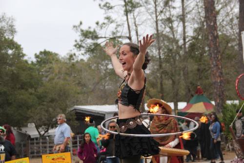 A performer hula hoops with fire, smiling and engaging a crowd at an outdoor festival surrounded by trees.