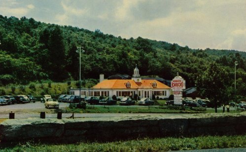 A vintage diner with an orange roof, surrounded by trees and parked cars, set against a hilly landscape.