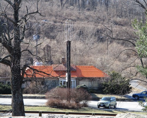 A small building with a red roof surrounded by bare trees and a parked car, set against a hilly background.