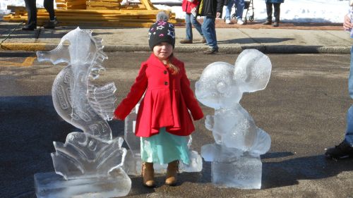A young girl in a red coat stands between two ice sculptures at a winter festival, with people and snow in the background.