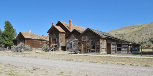 A row of rustic wooden buildings under a clear blue sky, set against a grassy landscape and distant hills.
