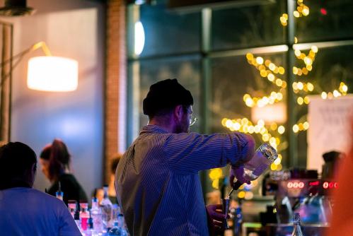 A bartender in a hat pours a drink at a lively bar with warm lighting and festive decorations in the background.