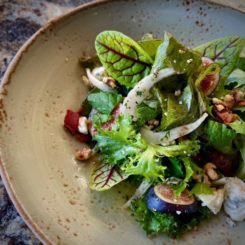 A fresh salad with mixed greens, figs, onions, and nuts served in a rustic bowl on a textured surface.