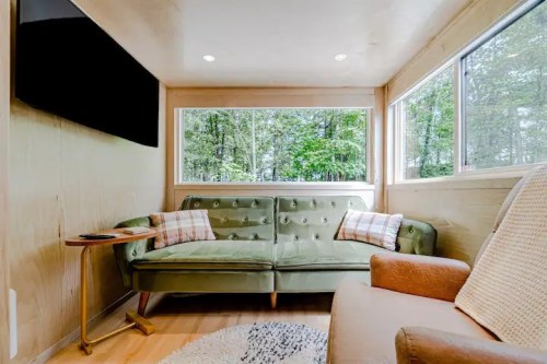 Cozy interior of a tiny home featuring a green sofa, armchair, and large windows with a view of trees outside.