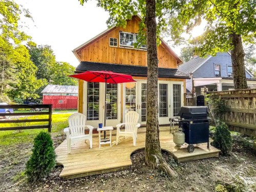 Cozy backyard with a wooden deck, two chairs, a red umbrella, and a grill, surrounded by trees and a rustic barn.