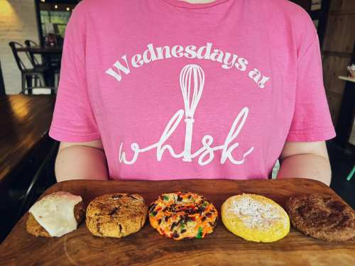 A person wearing a pink "Wednesdays at Whisk" shirt holds a wooden board with five colorful cookies.
