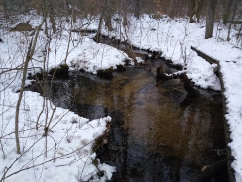 A serene stream flows through a snowy forest, with patches of snow on the banks and trees in the background.