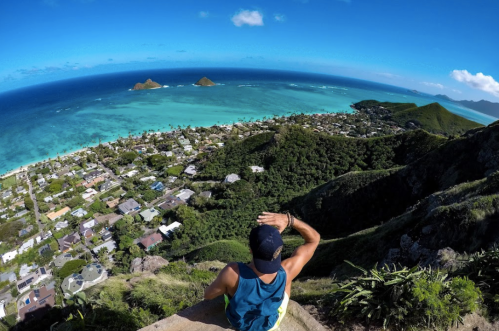 A person sits on a rocky ledge, overlooking a vibrant coastal landscape with islands and a clear blue sky.