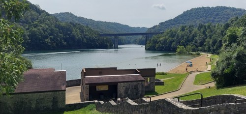Scenic view of a river surrounded by green hills, with a bridge in the background and a sandy area along the shore.