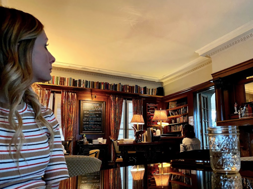 A woman with wavy hair sits in a cozy library, looking towards a bookshelf and a person at a desk in the background.