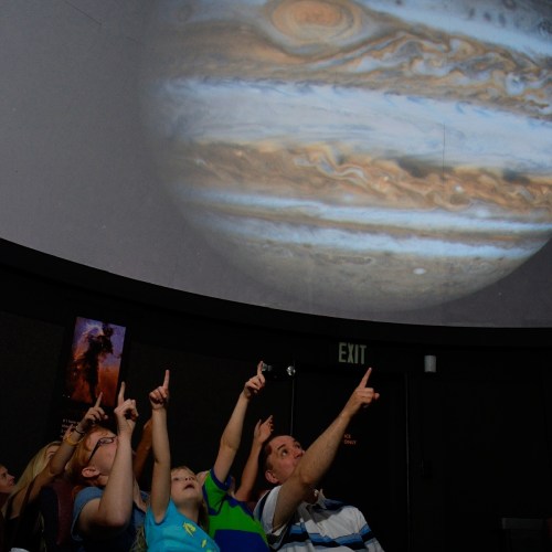 A group of people, including children, point at a large projection of Jupiter on a planetarium dome.