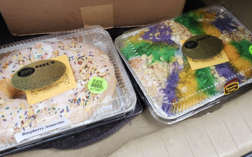 Two colorful king cakes in plastic containers, one raspberry amaretto and the other decorated with green, yellow, and purple.