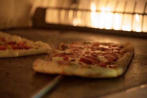 Two pizzas baking in a wood-fired oven, with one featuring pepperoni and melted cheese. Warm, glowing flames in the background.