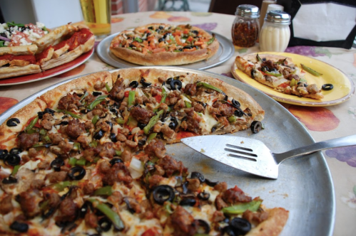 A variety of pizzas on a table, featuring toppings like sausage, olives, peppers, and a side of condiments.
