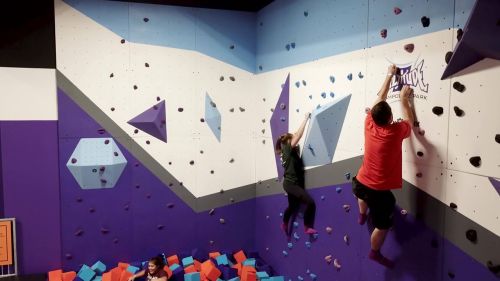 Two climbers scale a colorful bouldering wall, while a third person rests on a pile of foam blocks below.