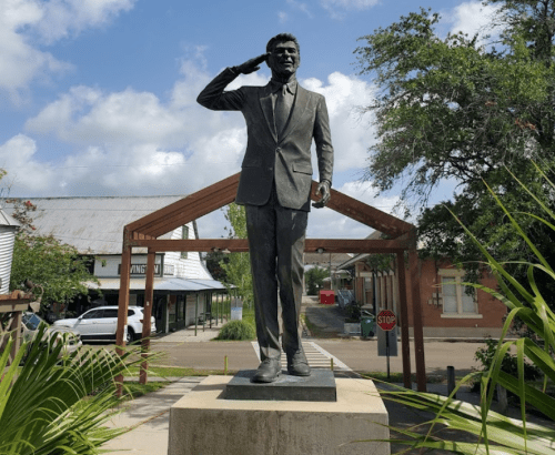 A bronze statue of a man saluting, set in a park with trees and buildings in the background.
