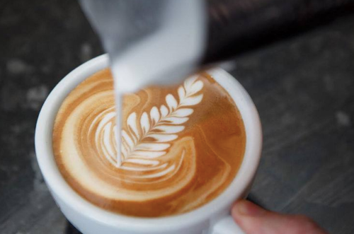 A close-up of a latte with intricate leaf latte art being poured from a milk jug.