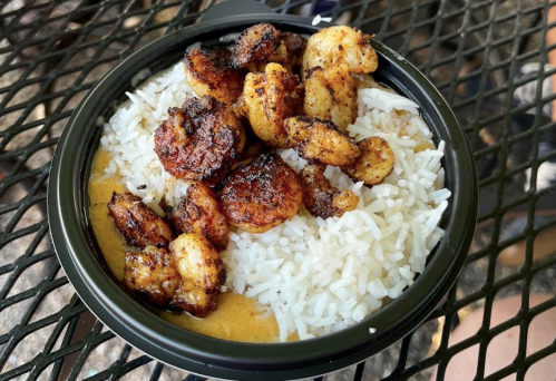A bowl of rice topped with grilled shrimp, served on a black container, resting on a metal table.